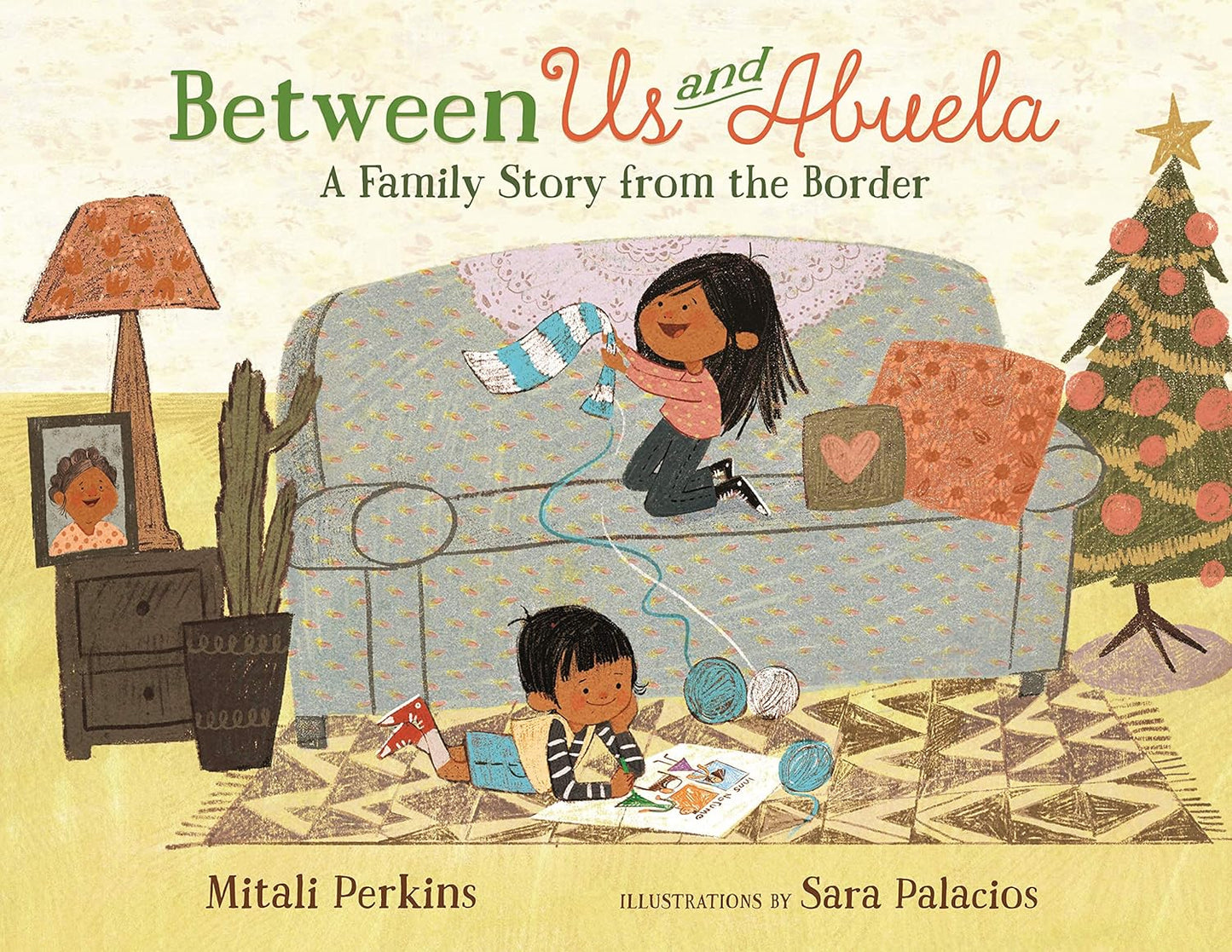 Cozy living room scene filled with warmth and love: a couch, a woman knitting, and a child playing with yarn beneath the glowing Christmas tree. Framed family photos and a lamp add charm. The book Between Us and Abuela: A Family Story from the Border by Mitali Perkins rests nearby.