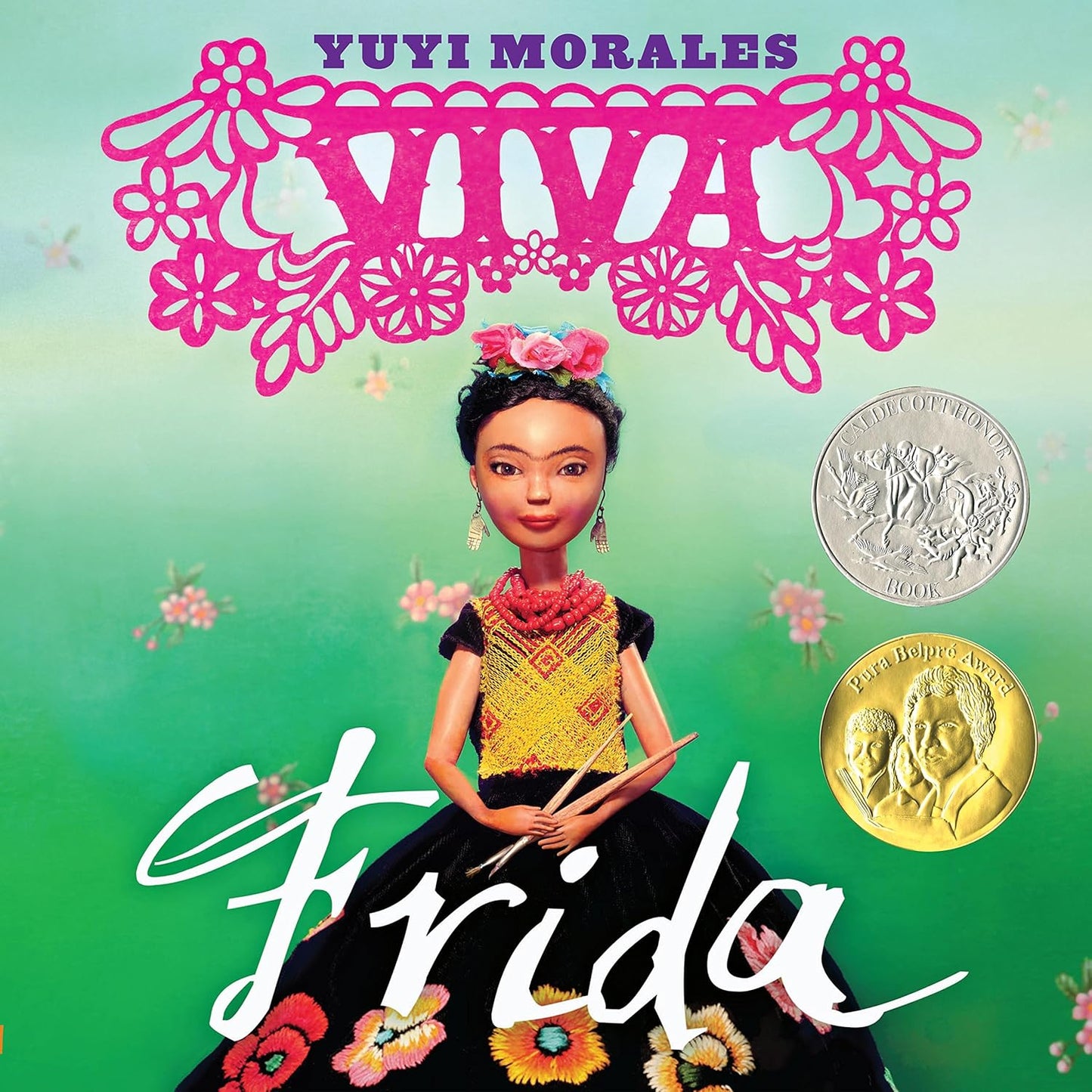The cover of Viva Frida features a doll-like figure resembling Frida Kahlo with flowers in her hair, against a green backdrop and pink cut-out designs, showcasing two award medals on the right.